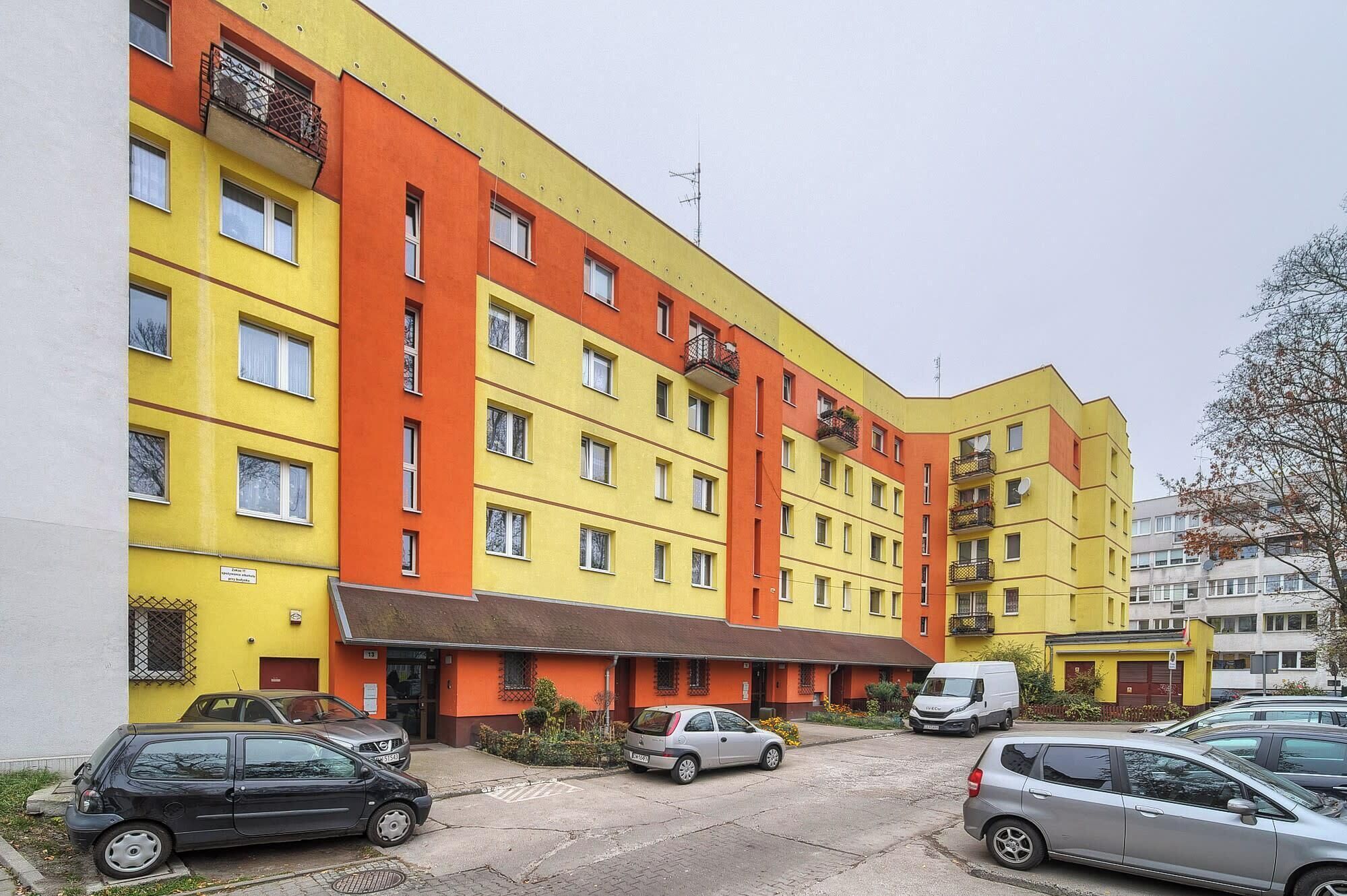 Um edifício de vários andares com fachada amarela e laranja, várias varandas e um estacionamento com carros.