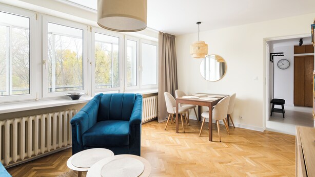 Una sala de estar moderna con un sillón azul, una mesa de comedor con sillas, un ventanal con cortinas, una lámpara colgante y un radiador.