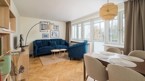 Una sala de estar moderna con un sofá azul, una mesa de comedor con sillas, una estantería y un ventanal con cortinas.