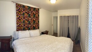 A bedroom with a large bed, a wooden nightstand, a colorful tapestry, and a door with curtains.