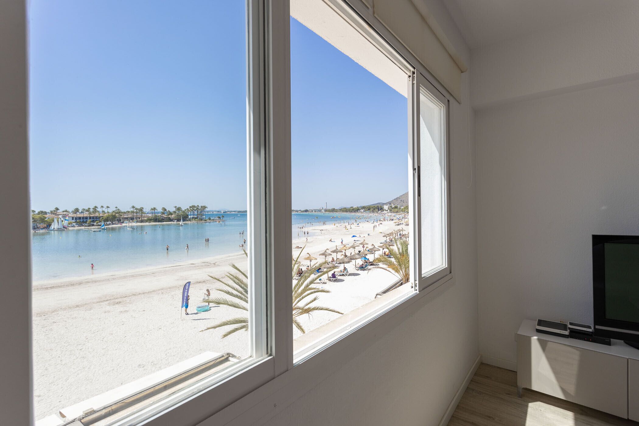 Una habitación con un amplio ventanal con vistas a una playa, un televisor sobre un mueble blanco y una mesita.