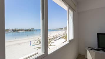 Una habitación con un amplio ventanal con vistas a una playa, un televisor sobre un mueble blanco y una mesita.