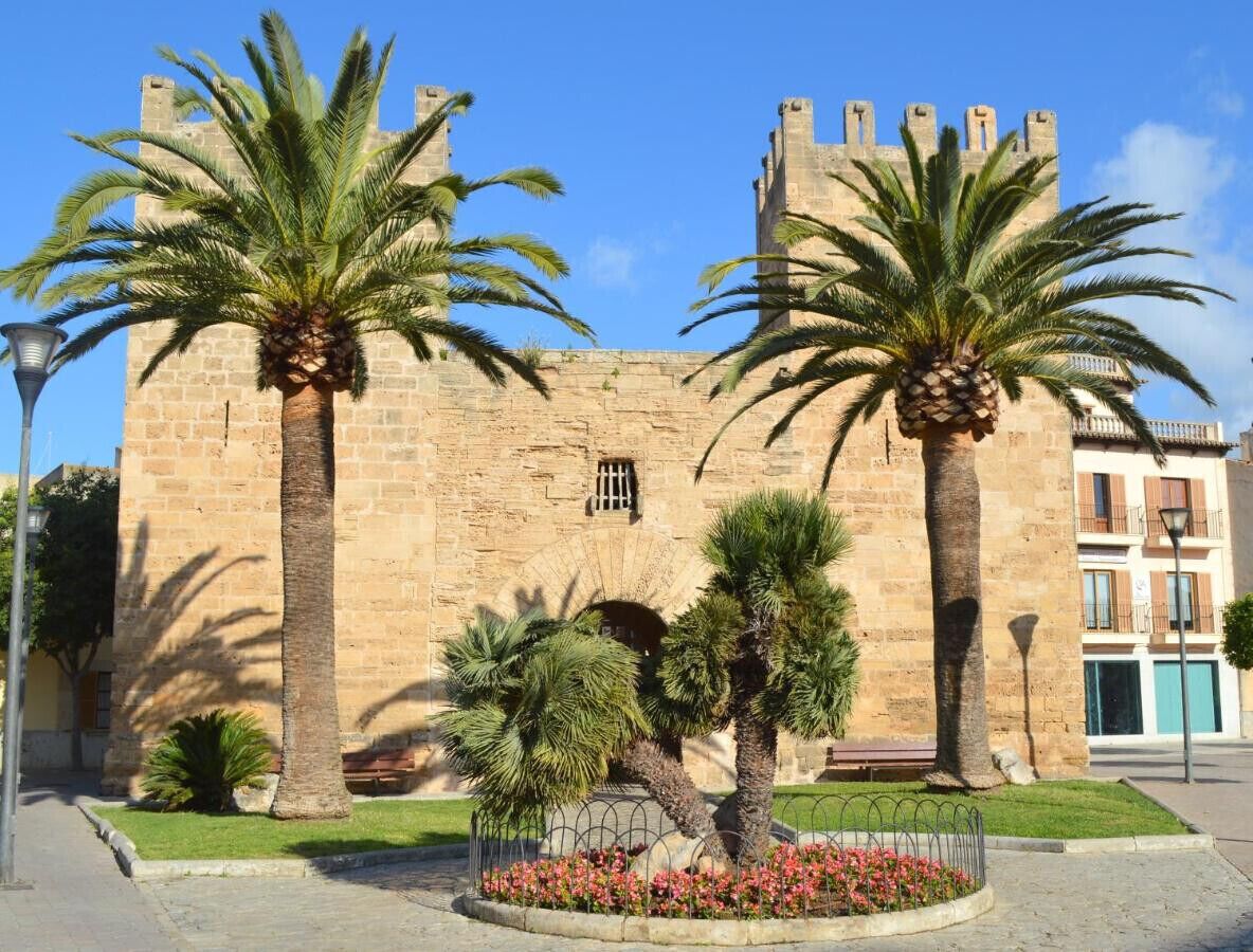 Un edificio histórico de piedra con palmeras y un jardín de flores al frente.