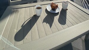 A deck with two Adirondack chairs, a table with a muffin and a cup of coffee, and a yellow door.