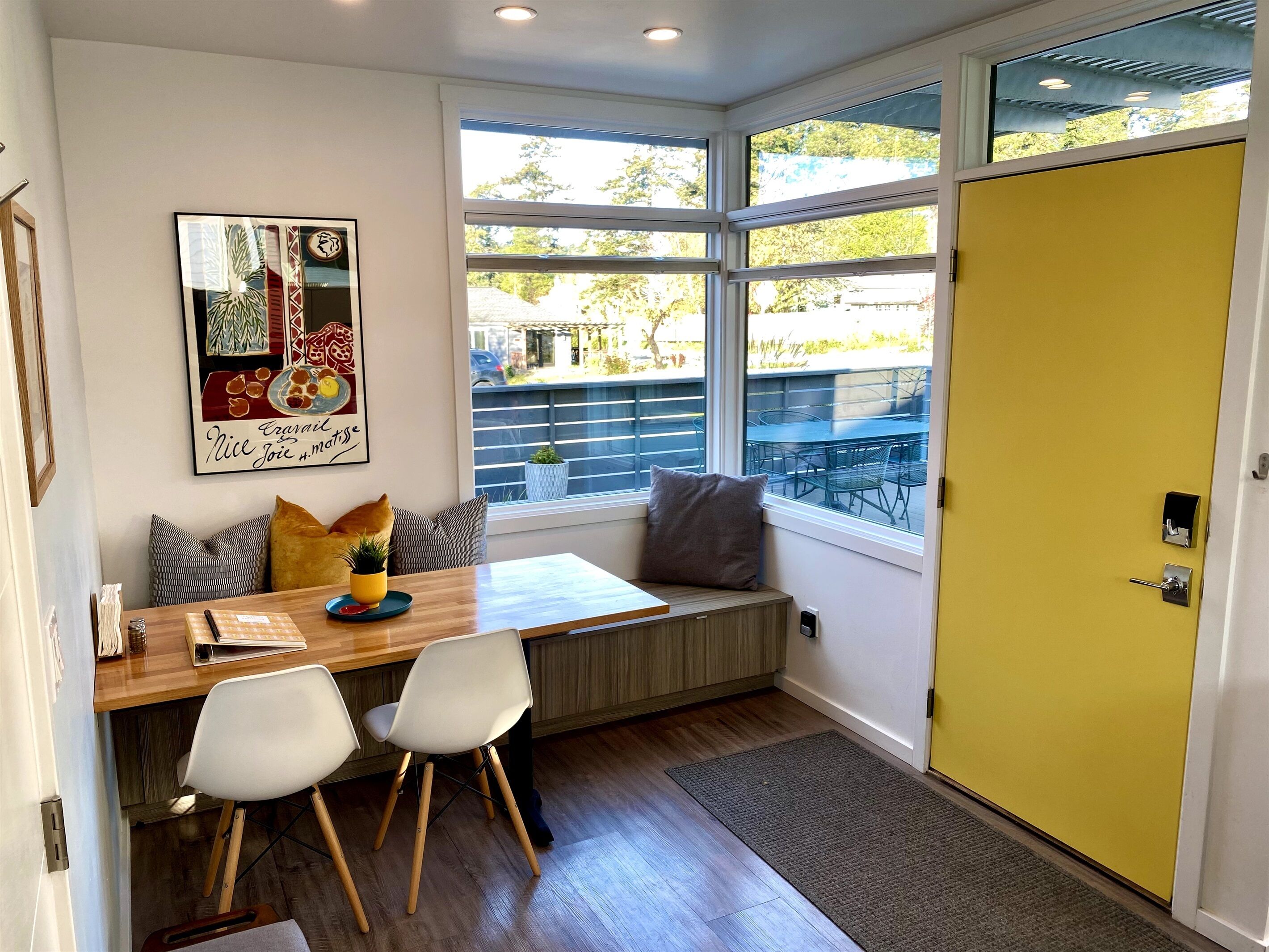 A modern kitchen with a wooden table, two white chairs, a bench with cushions, a large window, and a yellow door.