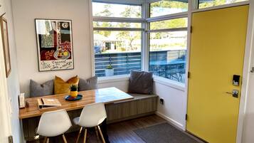 A modern kitchen with a wooden table, two white chairs, a bench with cushions, a large window, and a yellow door.