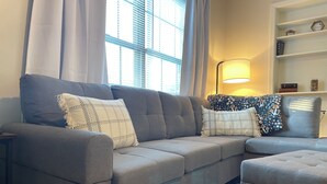 A modern living room with a grey sofa, patterned cushions, a side table, a lamp, and a bookshelf.