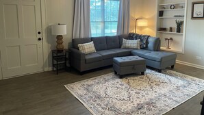 A living room with a gray sectional sofa, an ottoman, a patterned rug, a ceiling fan, and a window with curtains.