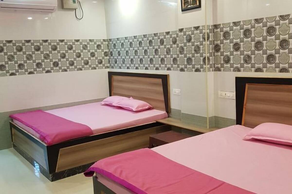 A hotel room with two beds, each with a wooden headboard and pink bedding. The room has a tiled backsplash and a small bedside table.