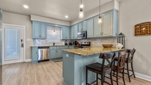 Une cuisine avec des meubles de couleur bleu clair, un îlot central doté de tabourets, des appareils en acier inoxydable et une fenêtre avec des stores.