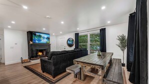A modern living room with a fireplace, a large TV, and a wooden dining table. - Cozy Ranch by the Lake w/Hot Tub in the Mountains (Albrightsville)