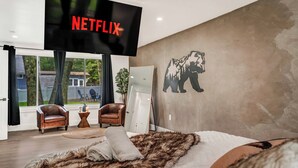 A modern bedroom with a large bed, a mounted TV displaying 'NETFLIX', a bear mural, and a view of the outdoors. - Cozy Ranch by the Lake w/Hot Tub in the Mountains (Albrightsville)