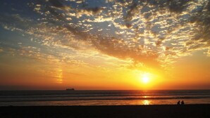 A sunset over the ocean with a ship in the distance and silhouettes of people on the beach.