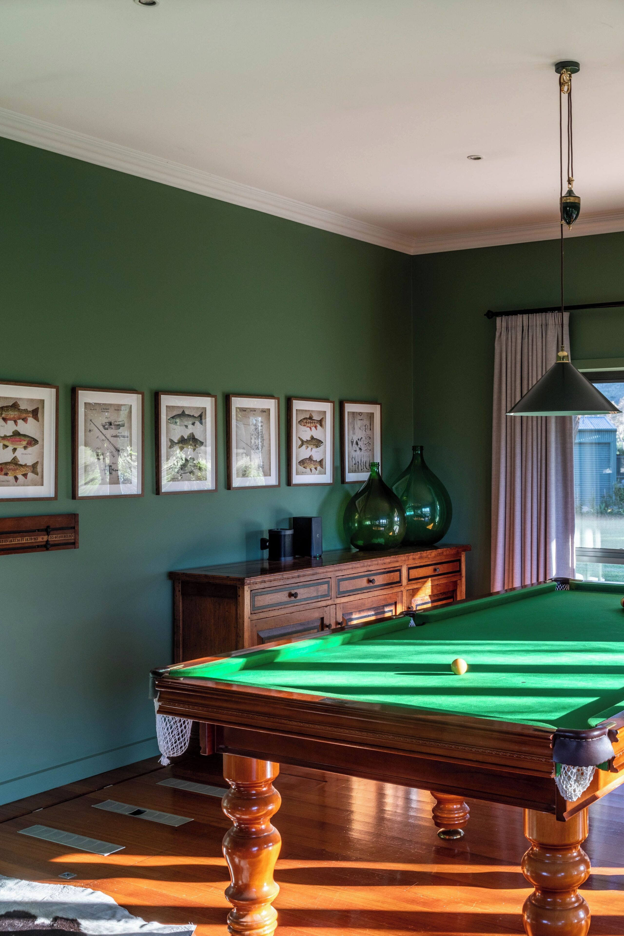 Una mesa de billar en una habitación con paredes verdes, cuadros de peces enmarcados y un mueble de madera.