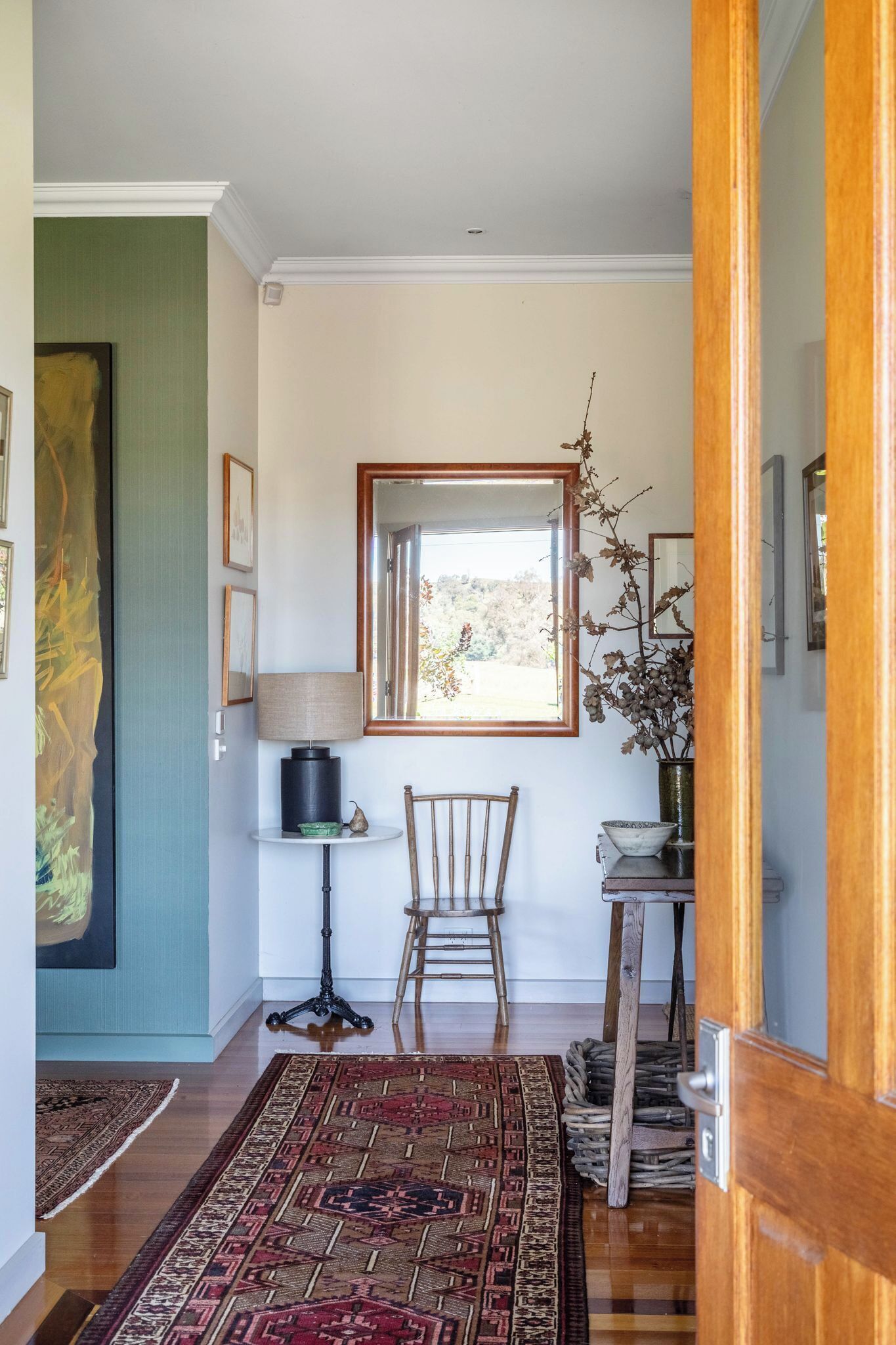 A hallway with a patterned rug, a chair, a lamp, and a mirror reflecting a view of trees.