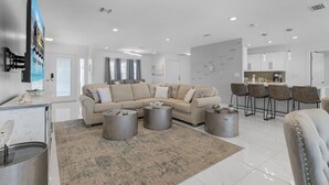 A modern living room with a beige sectional sofa, a coffee table, and a dining area with bar stools. - IMPECCABLE! - Amazing sunsets, Dock, Heated Pool - Sunsational Sunsets - Roelens (Cape Coral)