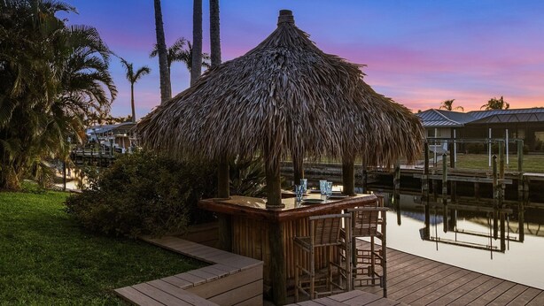 A thatched-roof bar area with wooden furniture by a calm water body, surrounded by greenery and palm trees. - IMPECCABLE! - Amazing sunsets, Dock, Heated Pool - Sunsational Sunsets - Roelens (Cape Coral)