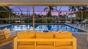A poolside area with a sofa and lounge chairs, a clear view of a swimming pool, and a sunset in the background. - IMPECCABLE! - Amazing sunsets, Dock, Heated Pool - Sunsational Sunsets - Roelens (Cape Coral)
