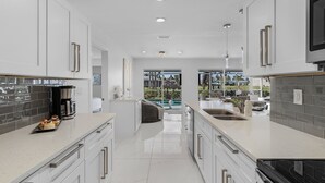 A modern kitchen with white cabinetry, a large island, and a view to an outdoor pool area. - IMPECCABLE! - Amazing sunsets, Dock, Heated Pool - Sunsational Sunsets - Roelens (Cape Coral)