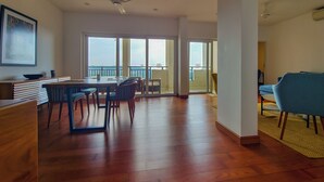 A spacious living area with a wooden floor, a dining table with chairs, a balcony with a view, and a ceiling fan.