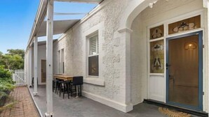 A covered outdoor seating area with a table and chairs, a white textured wall, and a blue door with glass panels. - Wanderlust 3Br Cottage Grange Illuminate Adelaide 2024 (Grange)