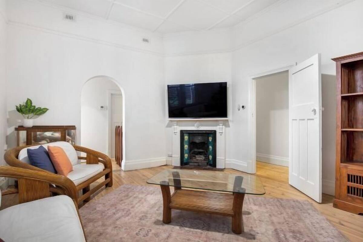 A living room with a glass-top coffee table, a sofa, a television mounted above a fireplace, and a wooden bookshelf.