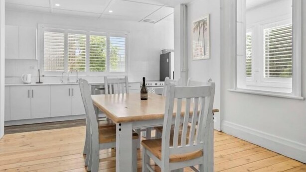 A modern kitchen with white cabinets, a wooden dining table, and a window with blinds. - Wanderlust 3Br Cottage Grange Illuminate Adelaide 2024 (Grange)