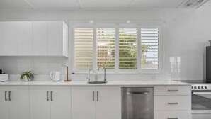 A modern kitchen with white cabinetry, a stainless steel dishwasher, and a window with blinds. - Wanderlust 3Br Cottage Grange Illuminate Adelaide 2024 (Grange)