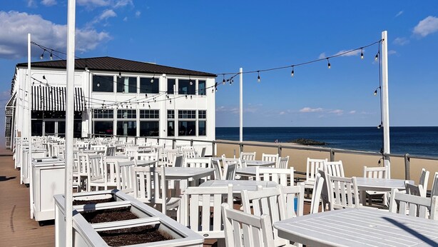 Ein Restaurant direkt am Strand mit Terrassensitzgelegenheiten, ein weißes Gebäude mit großen Fenstern und Blick auf den Ozean.