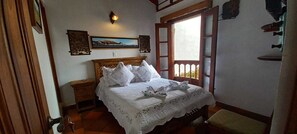 Une chambre à coucher avec un lit en bois, du linge de lit blanc, une table de chevet, une lampe fixée au mur et une fenêtre donnant sur un beau paysage.