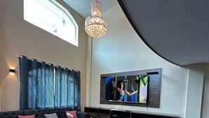 A modern living room with a large flat-screen TV mounted on the wall, a dark-colored sofa set, a wooden coffee table, and a chandelier. - Luxurious house near the airport - Tumbaco center (Quito)