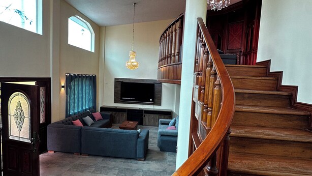 A spacious living room with a large sectional sofa, a flat-screen TV, and a fireplace. A wooden staircase with a curved railing leads to an upper level. - Luxurious house near the airport - Tumbaco center (Quito)