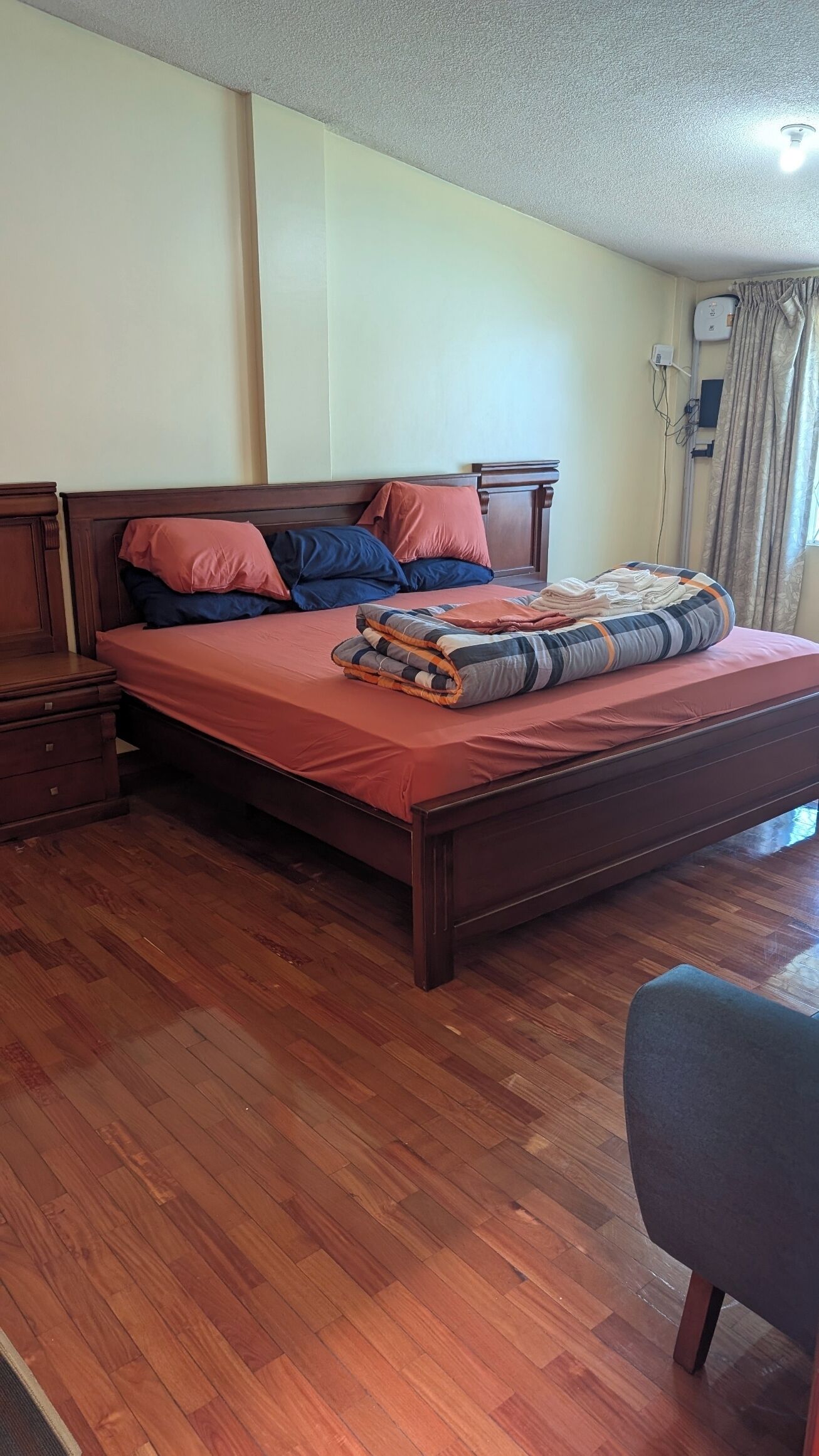 Un letto rifatto con cura, con una coperta rossa, una testiera in legno e numerosi cuscini. Accanto al letto, un comodino di legno, e una sedia in primo piano.