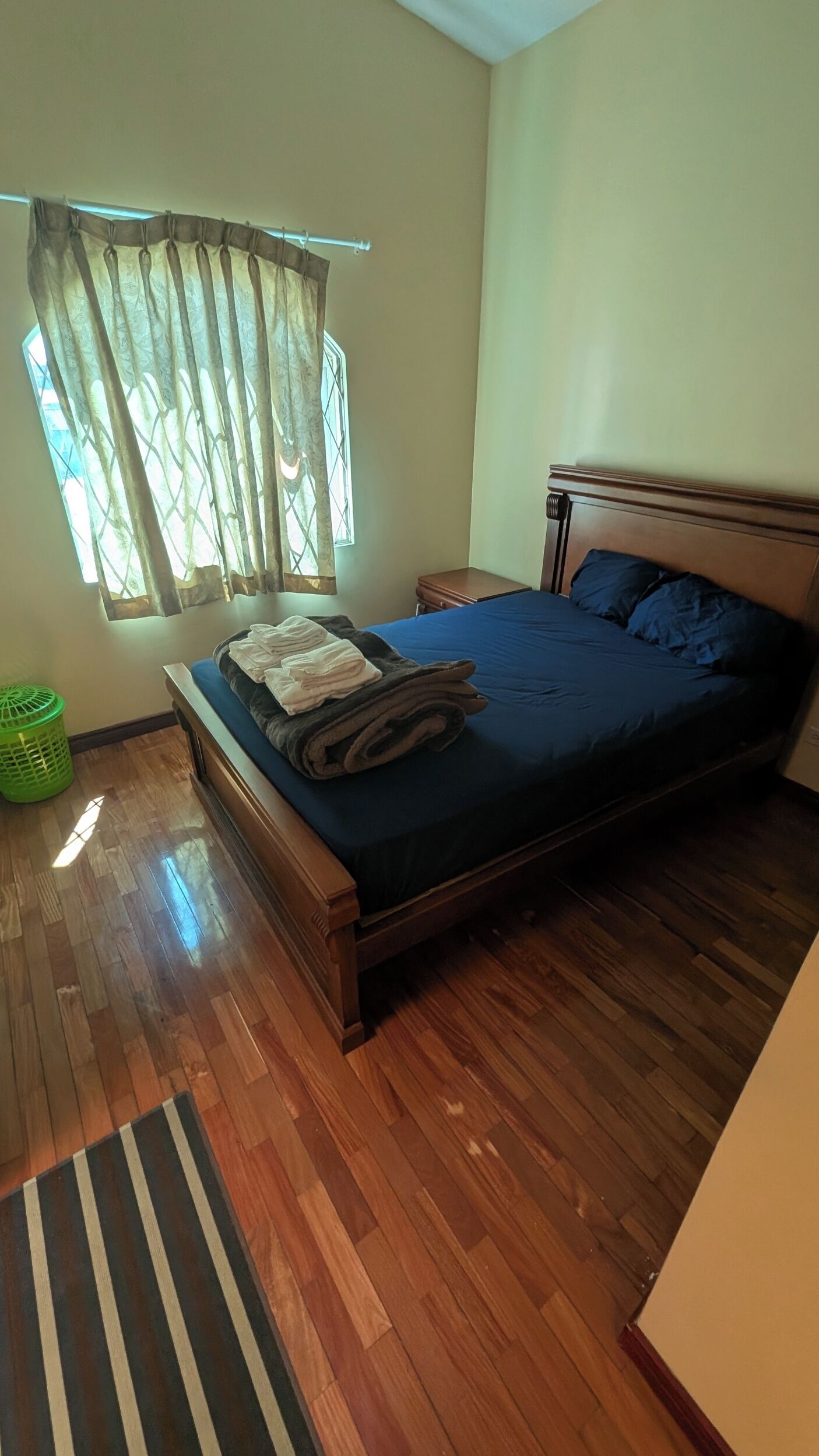 A bedroom with a wooden bed, a nightstand, a green laundry basket, and a window with curtains.