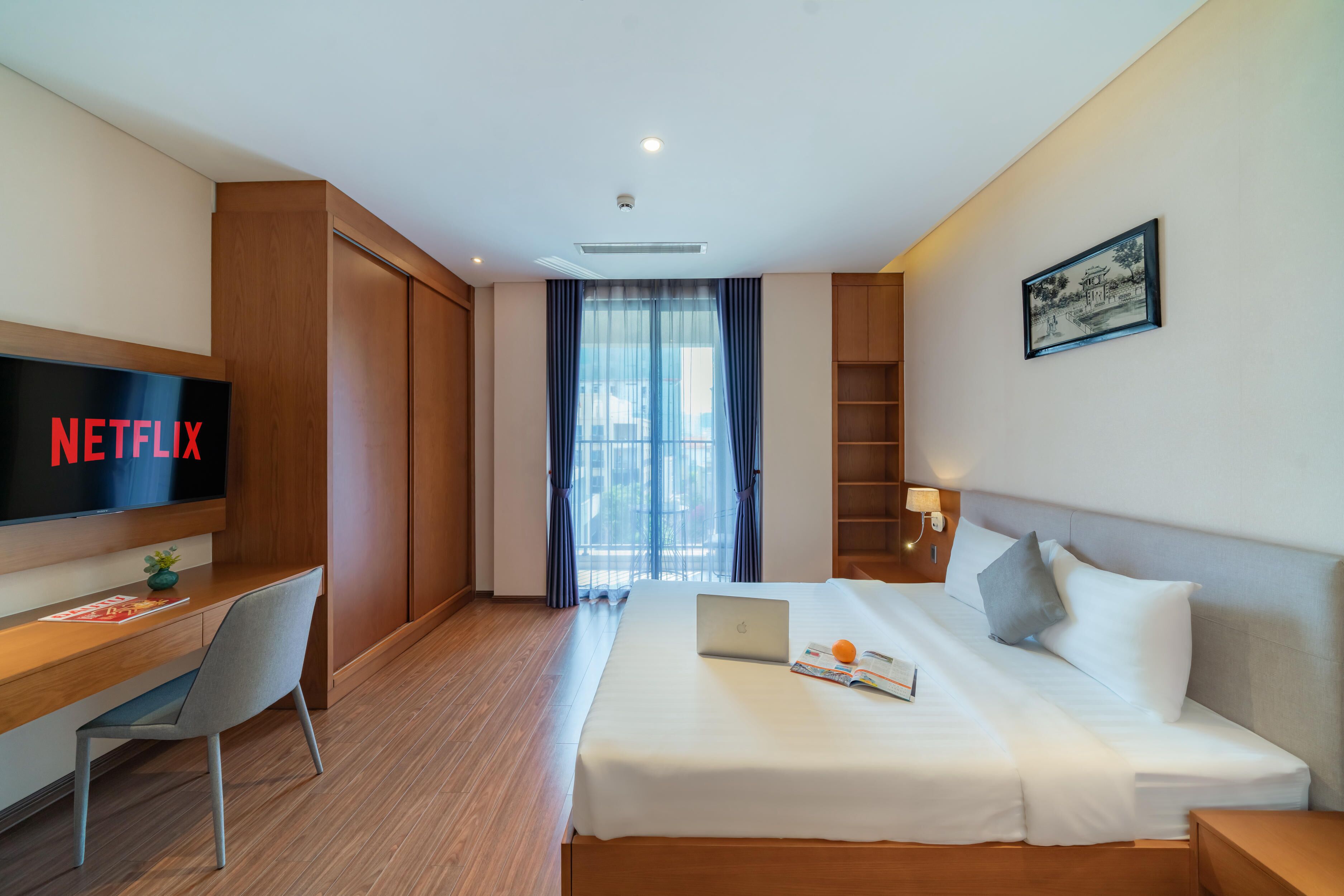 A hotel room with a bed, a desk with a laptop, a TV displaying 'NETFLIX', and a window with curtains.