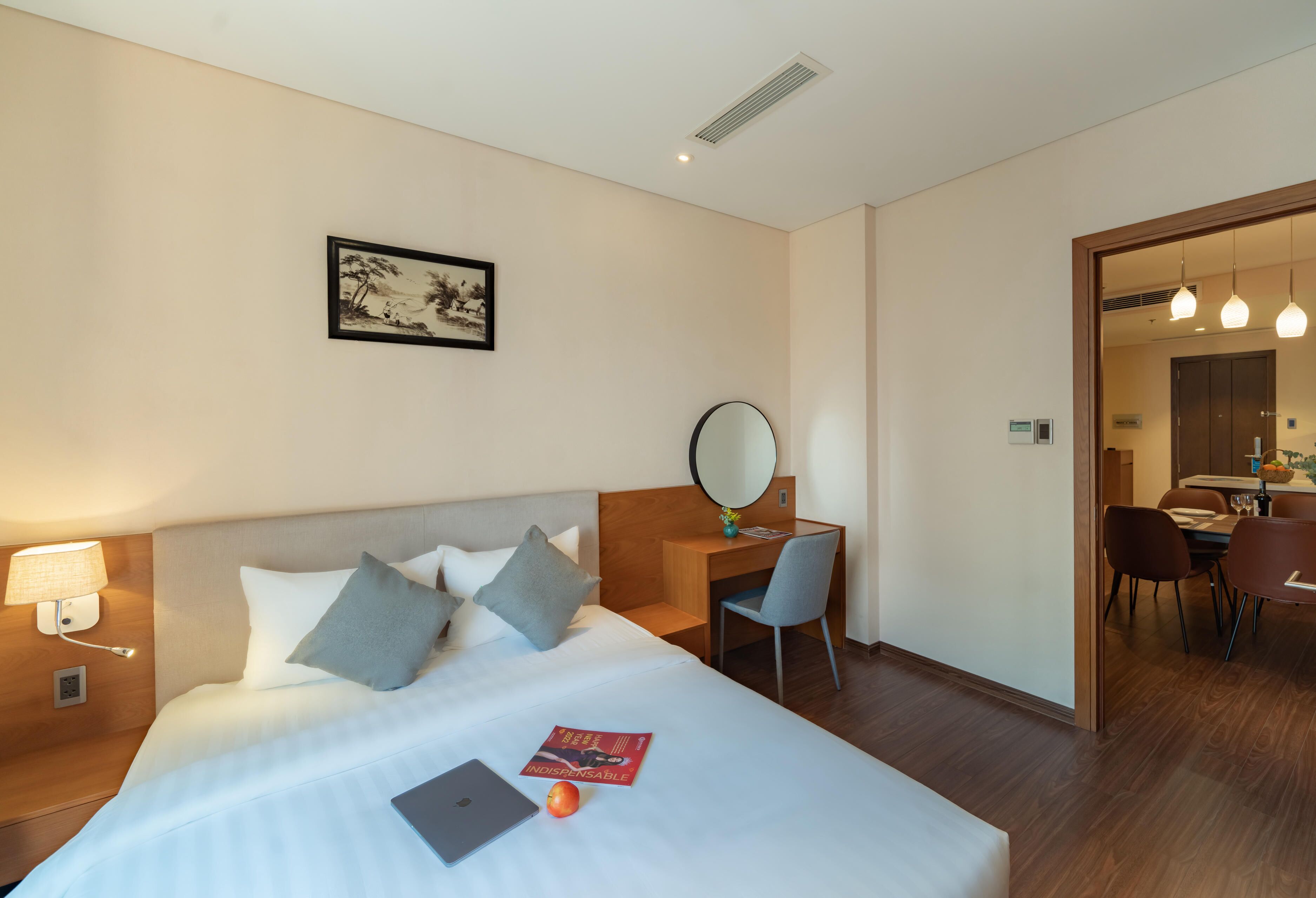 A hotel room with a bed, desk, chair, and a dining area visible through an open door.