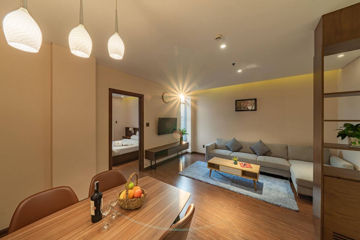 Ein modernes Wohnzimmer mit einer Couch, Couchtisch und Esstisch. Ein Fernseher ist an der Wand befestigt, ein Fenster mit Jalousien und eine Pflanze auf dem Fensterbrett.