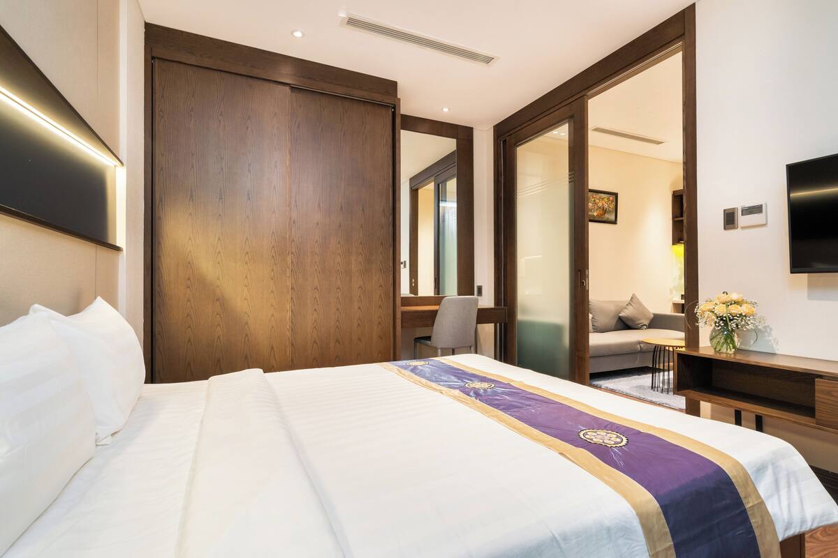 Ein Hotelzimmer mit einem großen Bett, einem Holzschrank, einem Schreibtisch, einem Sessel, einem Fernseher und einem kleinen Tisch mit Blumen.