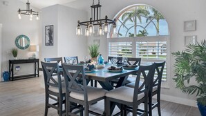 Un espace repas comprenant une grande table, des chaises et des luminaires suspendus. Une fenêtre offre une vue sur les palmiers, et un balcon est également présent.