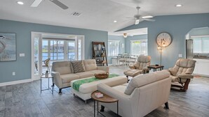 A spacious living room with a large sofa set, a coffee table, and a dining area with chairs. There are ceiling fans, a clock, and a view of the outdoors through large windows. - Charlotte County Villa 5872: Canal-Front Private Pool Oasis (Charlotte County)