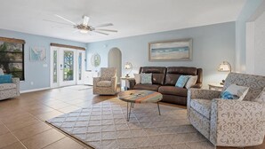 Ein geräumiges Wohnzimmer mit einer braunen Ledersofa, gemusterten Sessel und einem Couchtisch. An der Wand sind ein Deckenventilator und ein Gemälde zu sehen.