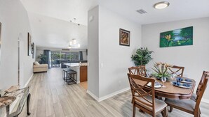 A spacious living area with a dining table set for four, a kitchen in the background, and a large window providing natural light.