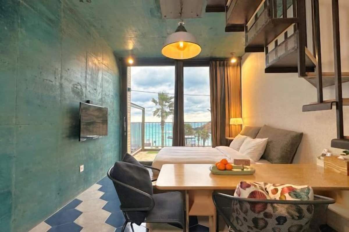 Une chambre d’hôtel comprenant un lit, un bureau avec une chaise, une table avec un panier de fruits, une télévision et un balcon avec vue sur la plage.