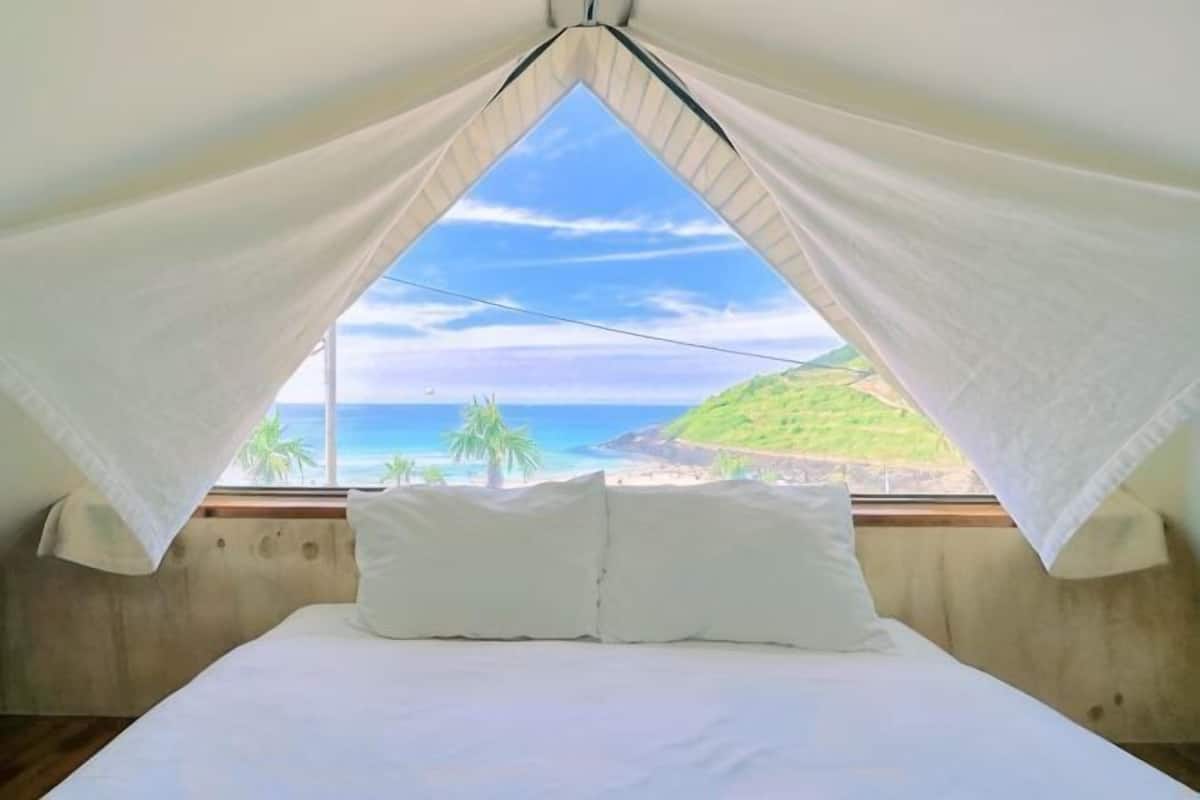Un lit avec du linge de lit blanc, une vue sur l’océan et une colline, et une chambre au décor simple.