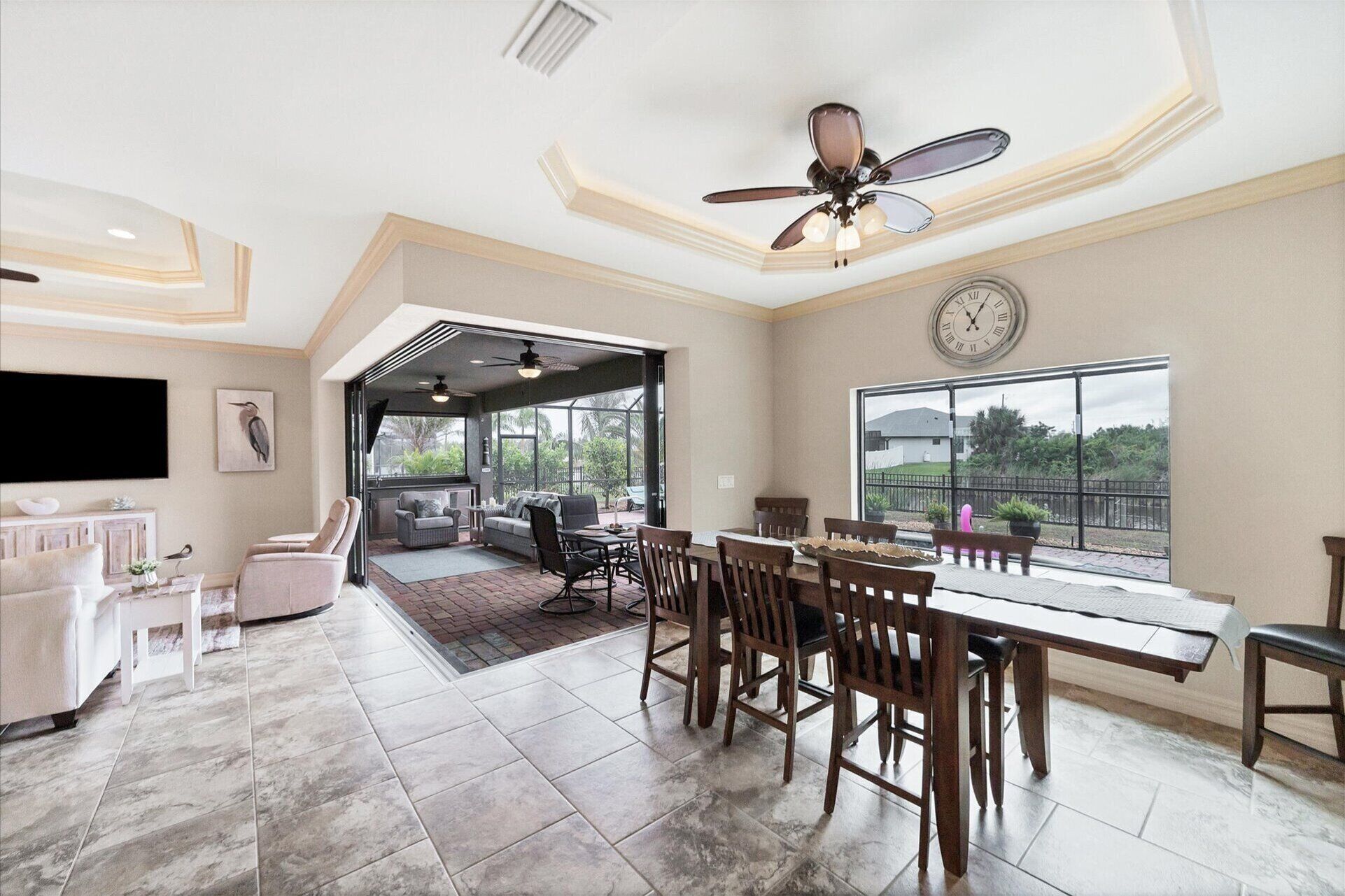 A spacious living area with a dining table, chairs, and a ceiling fan. There is a large window providing a view of the outdoors.