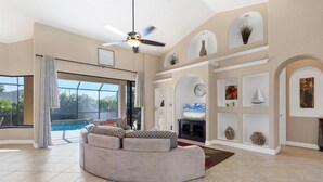 Un salón espacioso con un sofá curvo, un ventilador de techo, un televisor grande y una piscina visible a través de puertas de cristal.