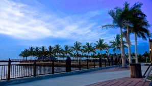 A waterfront promenade with a brick walkway, palm trees, and a metal railing.
