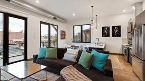 A modern apartment with a living room, dining area, and kitchen. There is a large window in the living room, a dining table with chairs, and a kitchen with a refrigerator and oven. - Sunrise Escape at Downtown Whitefish (Whitefish)