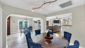 A spacious dining area with a round table and blue upholstered chairs, a kitchen with a large TV, and a ceiling fan. - Charming Charlotte County Villa 5884 with Private Pool Access (Charlotte County)