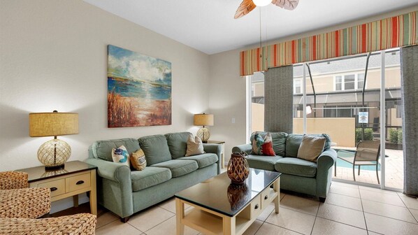 Ein Wohnzimmer mit zwei grünen Sofas, einem Glastisch und zwei Lampen. An der Wand hängt ein Gemälde und es gibt einen Blick auf einen Pool.